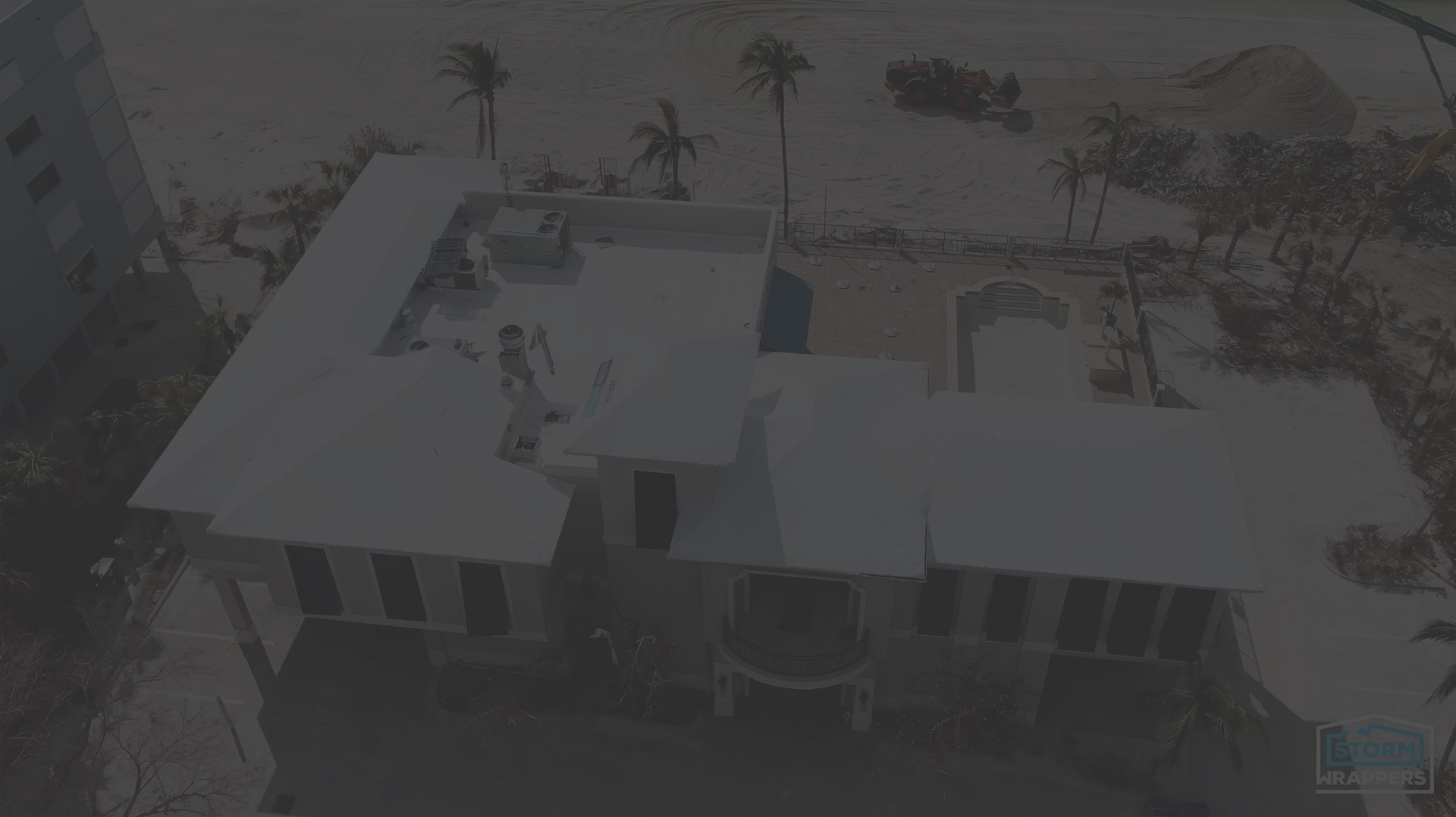 Aerial view of a white multi-story building under construction along a beach with palm trees and sand dunes in the background.