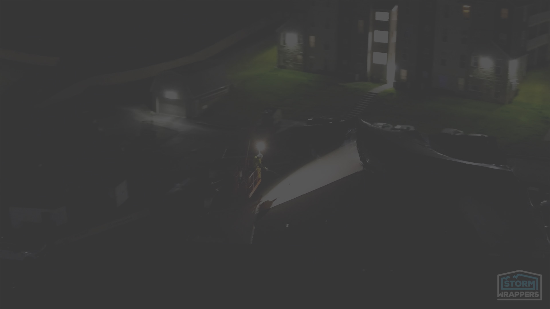 Night aerial view of a residential building at night with a worker on a lifted platform; Storm Wrappers logo in the bottom-right corner.