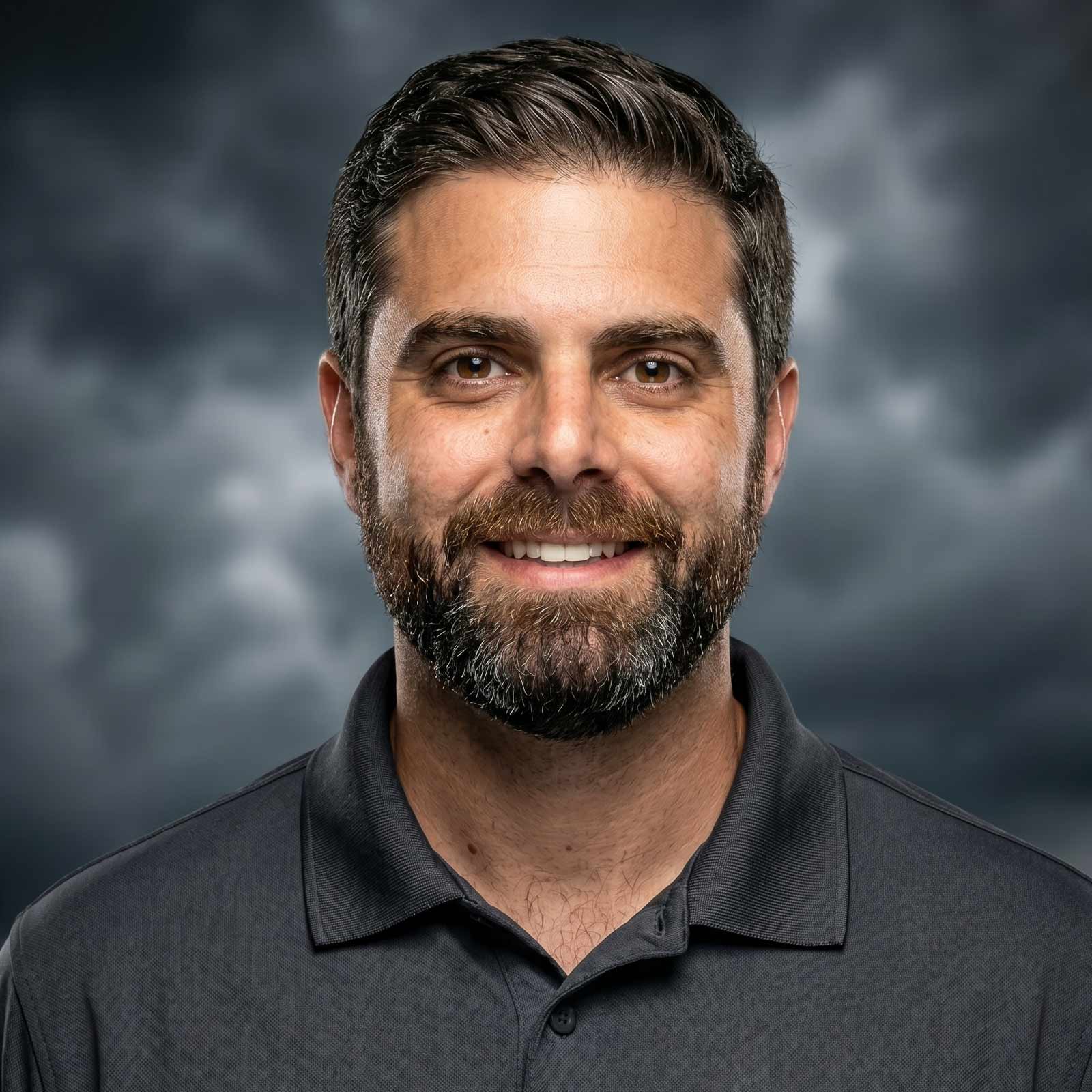 Close up portrait of a smiling man with dark hair and a beard wearing a black polo against a cloudy background