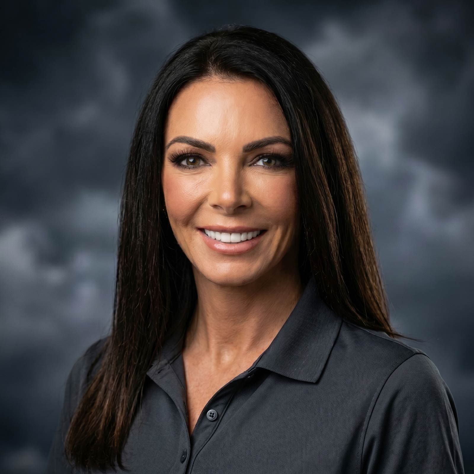 Portrait of a smiling woman with long dark hair and brown eyes wearing a dark collared shirt against a cloudy studio backdrop