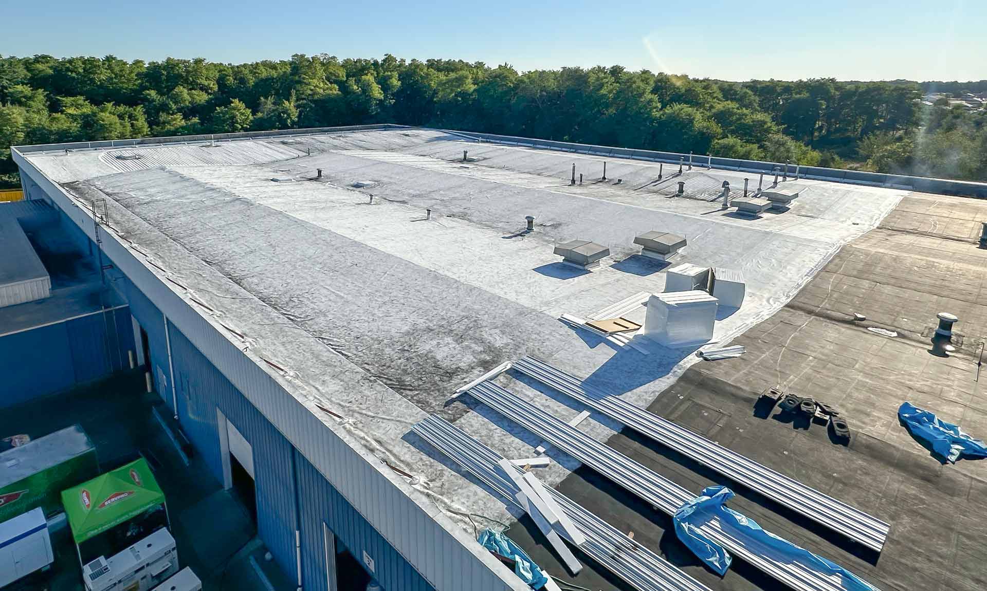 A flat industrial rooftop with weathered metal sheeting vents and scattered metal beams with trees visible beyond the edge of the building