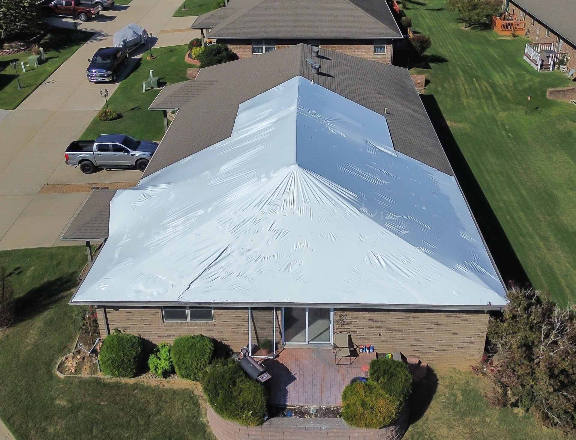 Aerial view of a suburban house with a large white tarp covering the roof with trucks in the driveway and well kept green lawns nearby