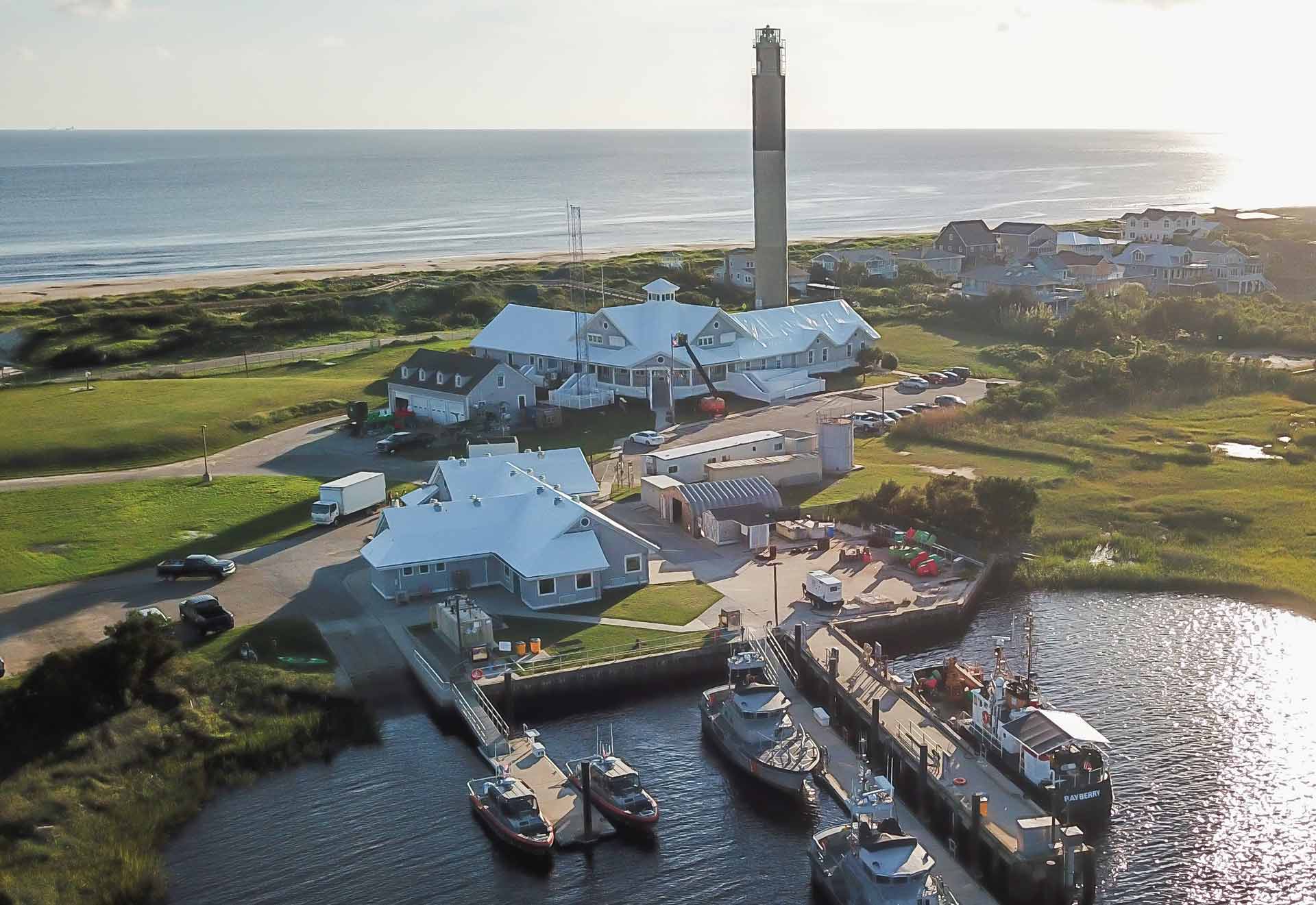 Aerial view of a coastal marina with white connected buildings and a tall lighthouse near the shore boats docked in the harbor