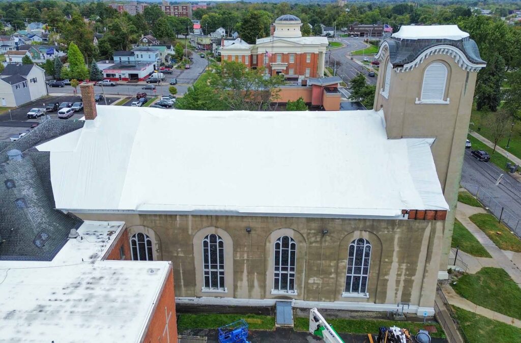 High angle view of a beige church with tall arched windows its roof covered in white tarps overlooking a small town with trees and streets below