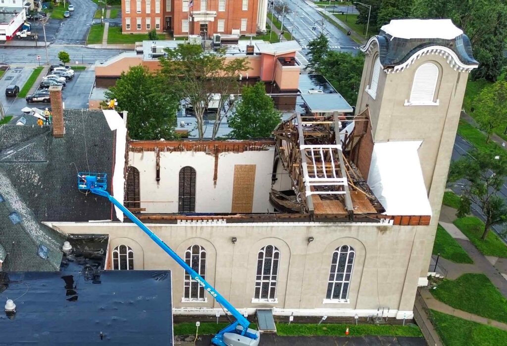 Aerial view of a tall beige building with a partially destroyed roof exposed wooden framework and a blue construction lift reaching toward the roof Parked cars and green trees surround the site