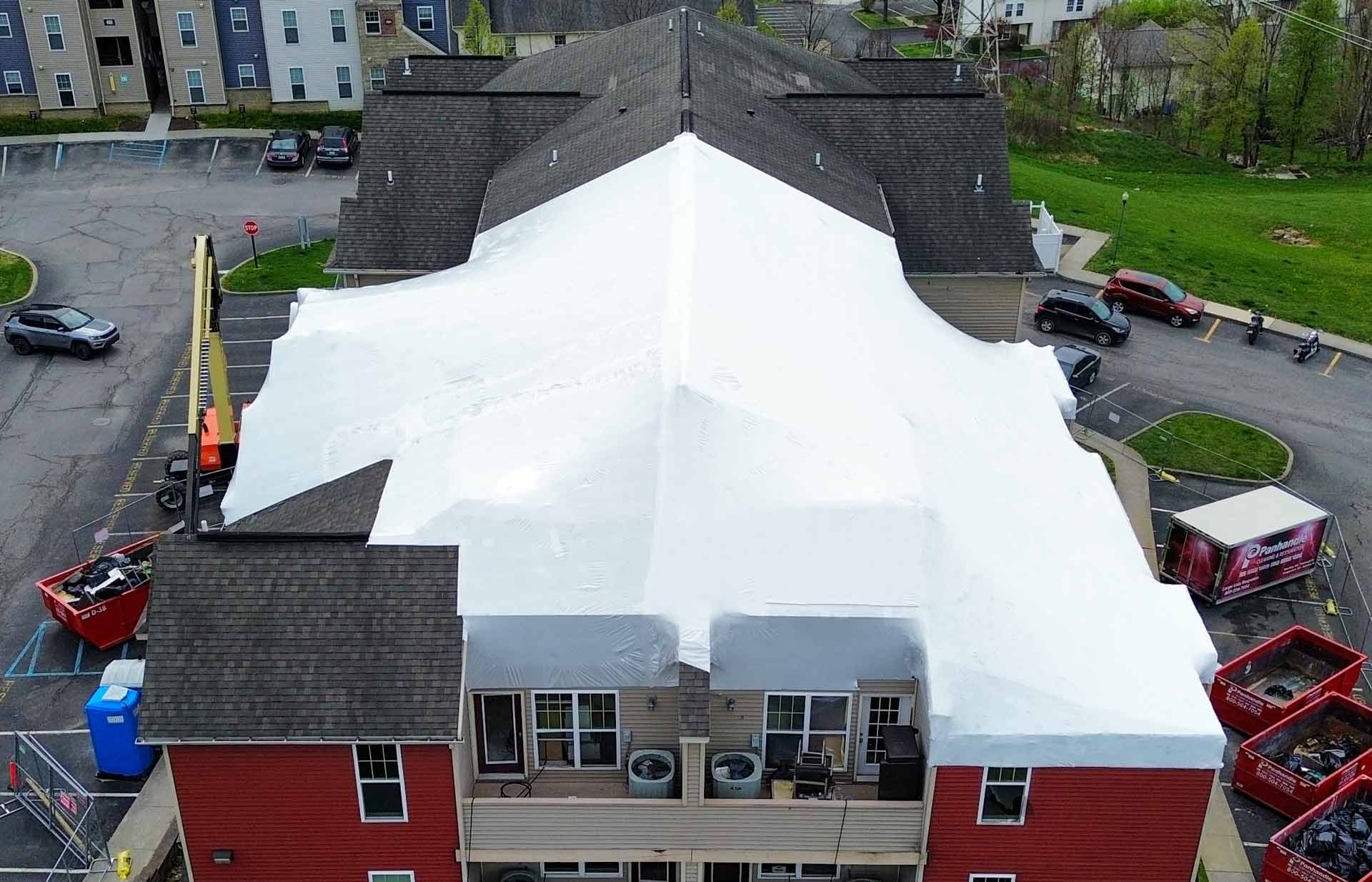 Aerial view of a red two story building covered by a large white canopytent over the roof with parked cars and dumpsters nearby in a parking lot