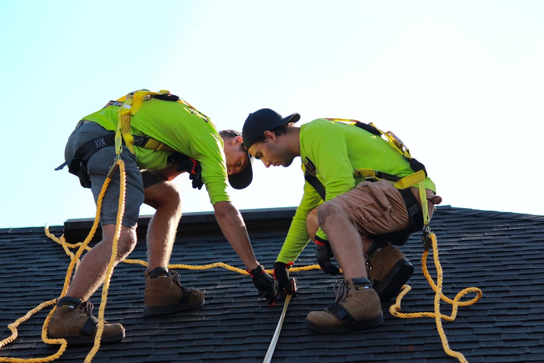 Two roof workers in bright green shirts and harnesses on a sloped roof, attaching a rope for fall protection.