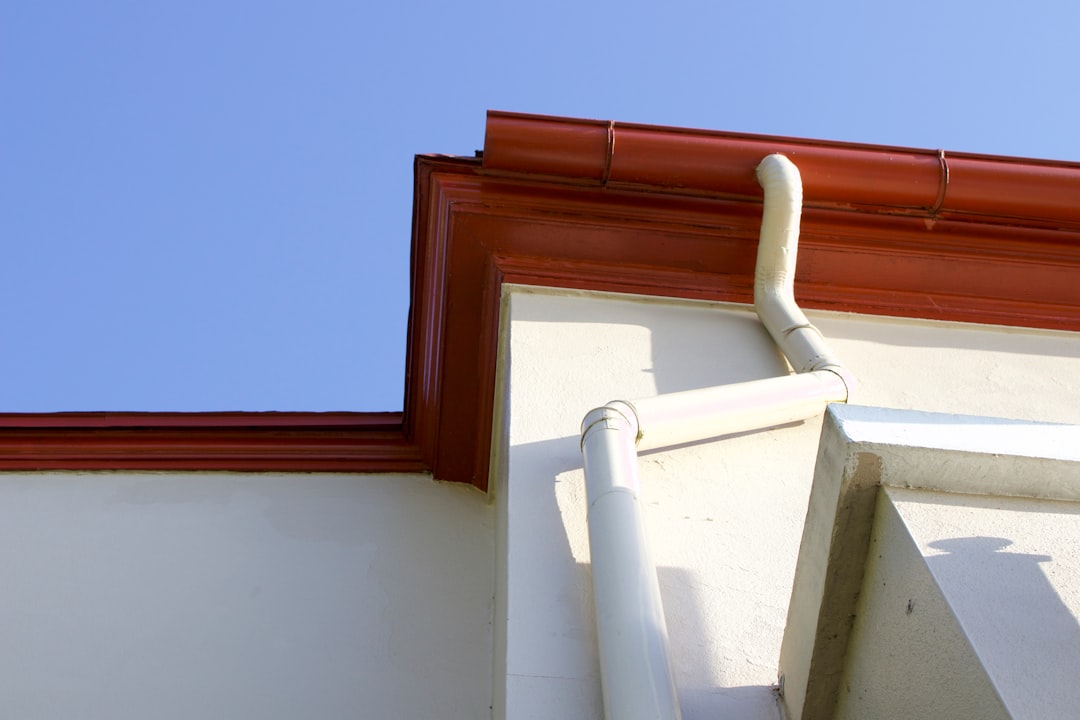 Close-up of white rain gutters and downspouts along a white building with a red metal roof trim against a blue sky.