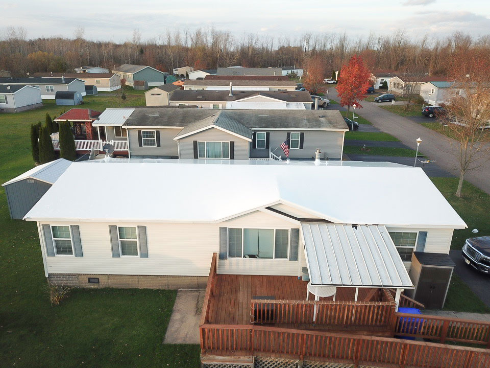 Drone view of a single story manufactured home with a white roof and wooden deck in a mobile home park suburban neighborhood fall trees in the background