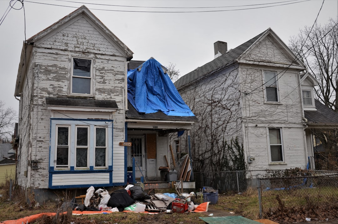 Two-story dilapidated white house with blue trim; blue tarp covers the roof, with a pile of debris and trash in the front yard.