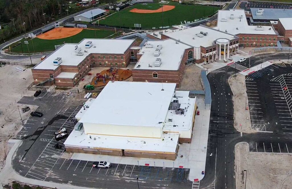 Aerial view of a school campus with brick buildings white roofs a playground baseball fields and surrounding parking lots and roads