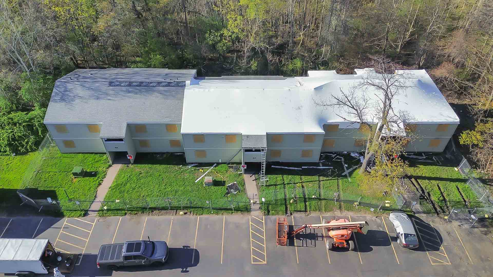 Aerial view of a long gray and white building complex under construction with a fenced yard trees behind and cars in a front parking lot