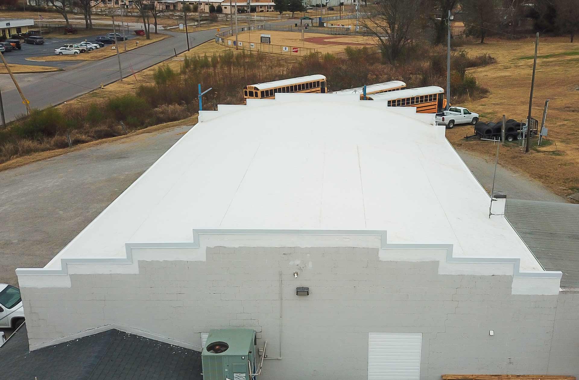 Aerial view of a white flat roofed building with decorative stepped edges three yellow school buses and some vehicles are parked behind it in a lot